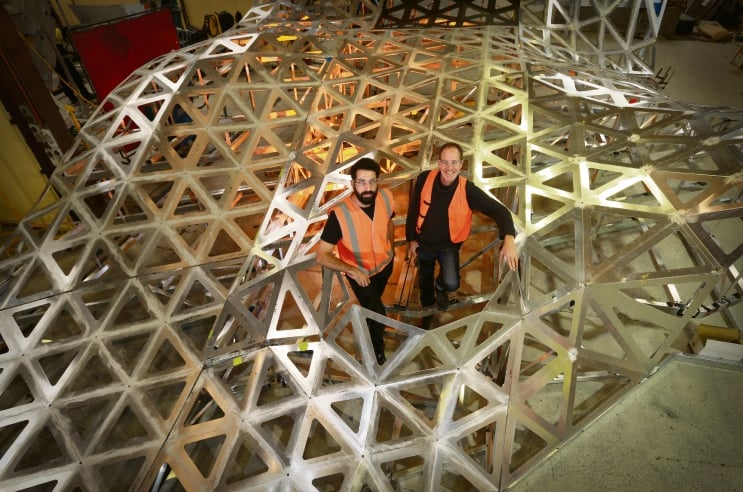 Rick Vermey and Daniel Giuffre in high visibility vests standing inside the triangulated aluminium frame of Anemoi on the workshop floor at Denmac