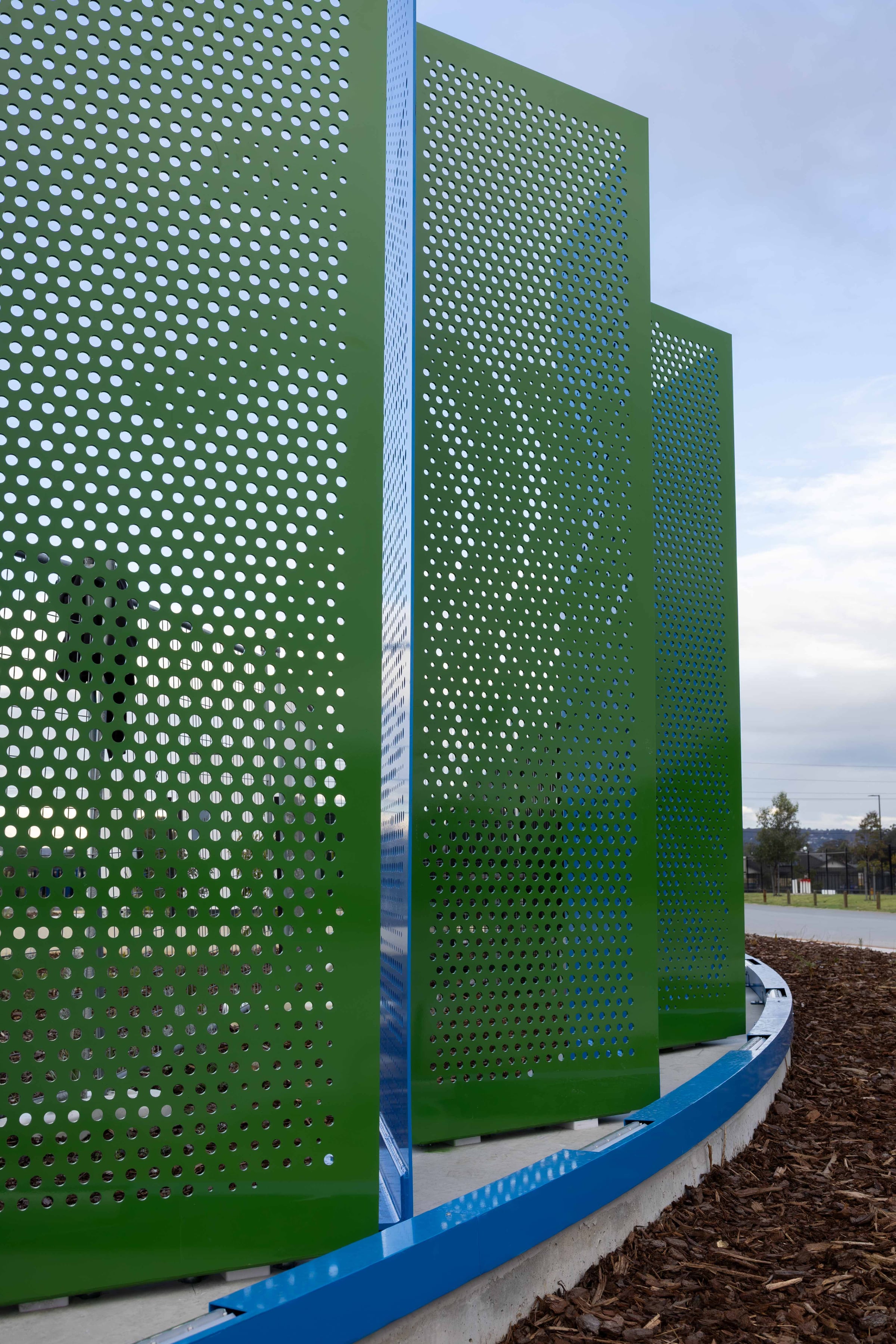 Green faces of the pleated steel panels catch the light above a curved blue plinth and fresh mulch.