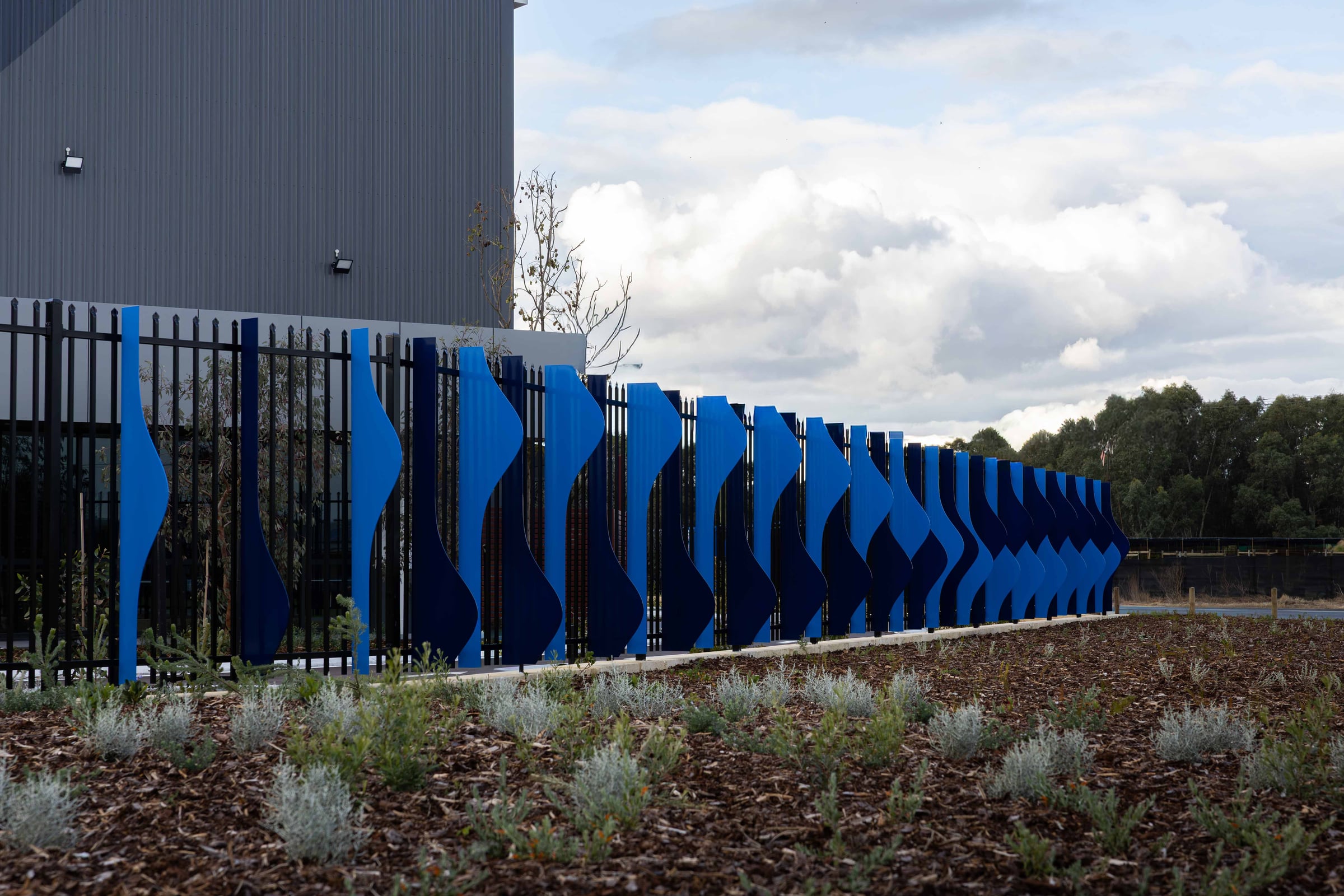Alternating pale and deep blue aluminium fins of Cascading Creeks line the warehouse fence in Hazelmere.