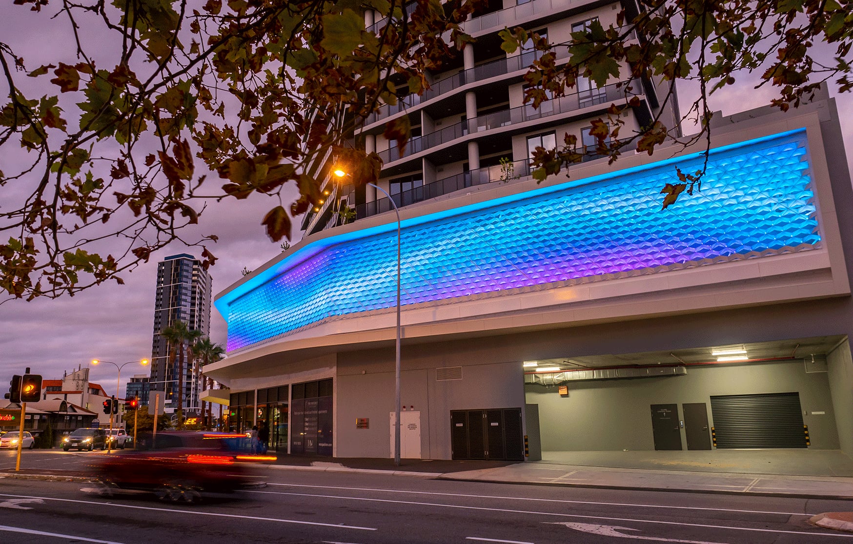 Dusk view of The Precinct apartments in Mt Pleasant, the folded facade glowing blue and violet above the street